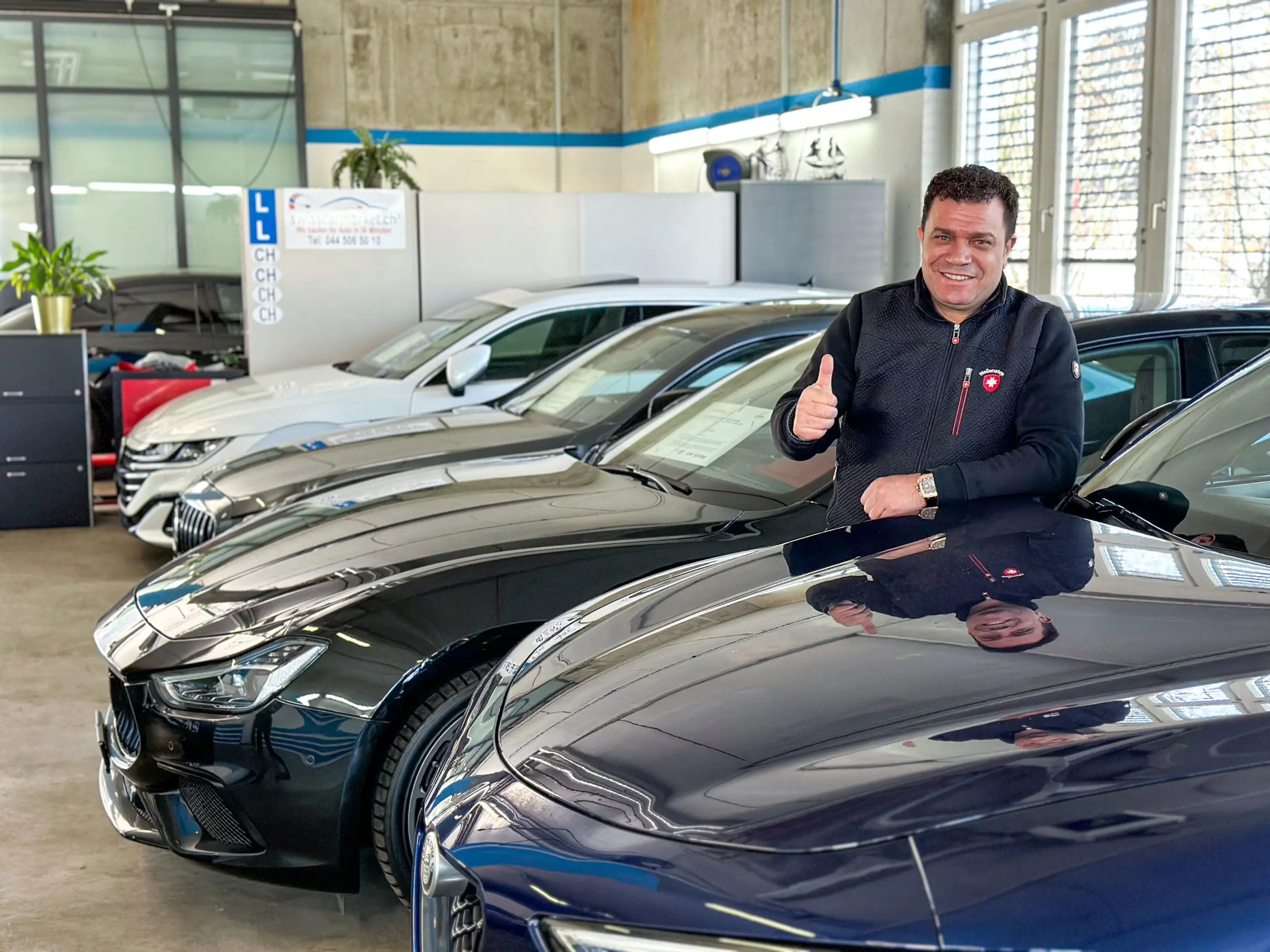 Ein lächelnder Mann im Showroom lehnt sich auf ein Auto und gibt einen Daumen hoch vor einer Reihe von Luxusfahrzeugen.